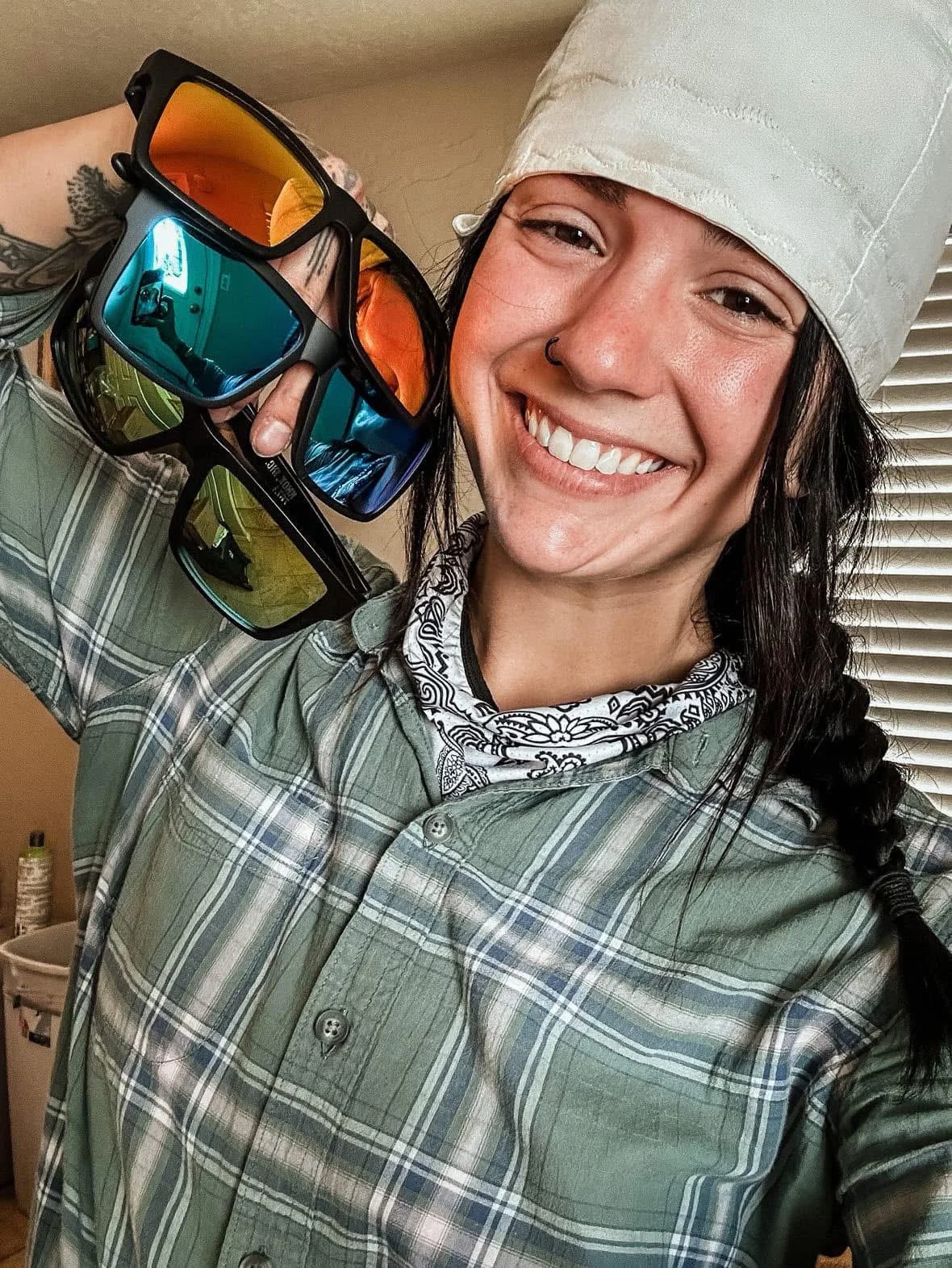 Tradeswoman wearing a welding cap and bandana holding Næxon Badger safety sunglasses with blue, orange, and green mirrored lenses